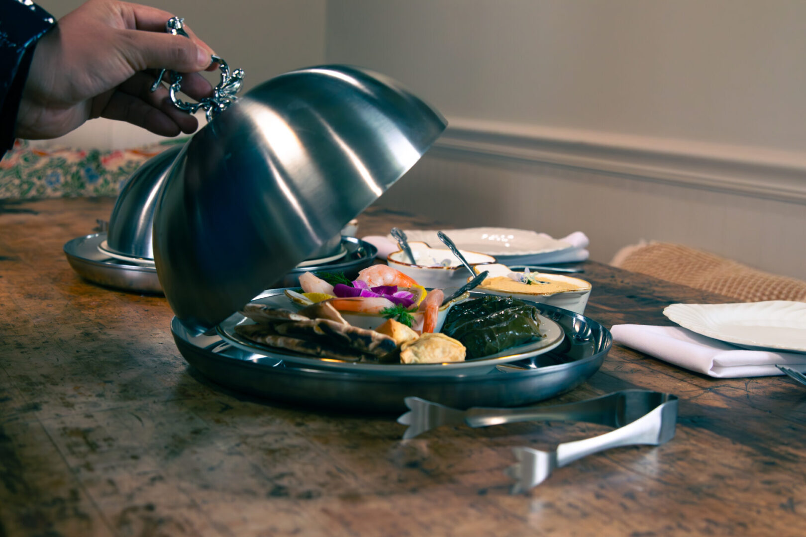 A hand lifting a cloche to reveal a gourmet dish on a plate.